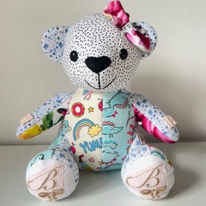 May include: A handmade teddy bear with a white polka dot head and a black heart-shaped nose. The bear has a pink bow and patches of colorful fabric with donuts, rainbows, and dinosaurs. The bear's feet are embroidered with a letter "B".