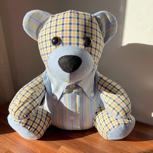 May include: A blue and yellow plaid teddy bear with button eyes and a blue and white striped shirt. The bear is sitting on a wooden surface.