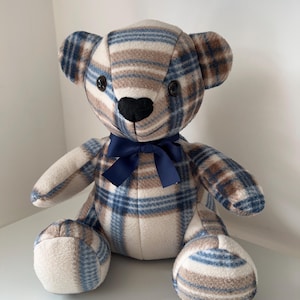 May include: A plush teddy bear with a plaid pattern in shades of blue, brown, and cream. The bear has black button eyes, a black nose, and a navy blue bow. The bear is sitting upright.