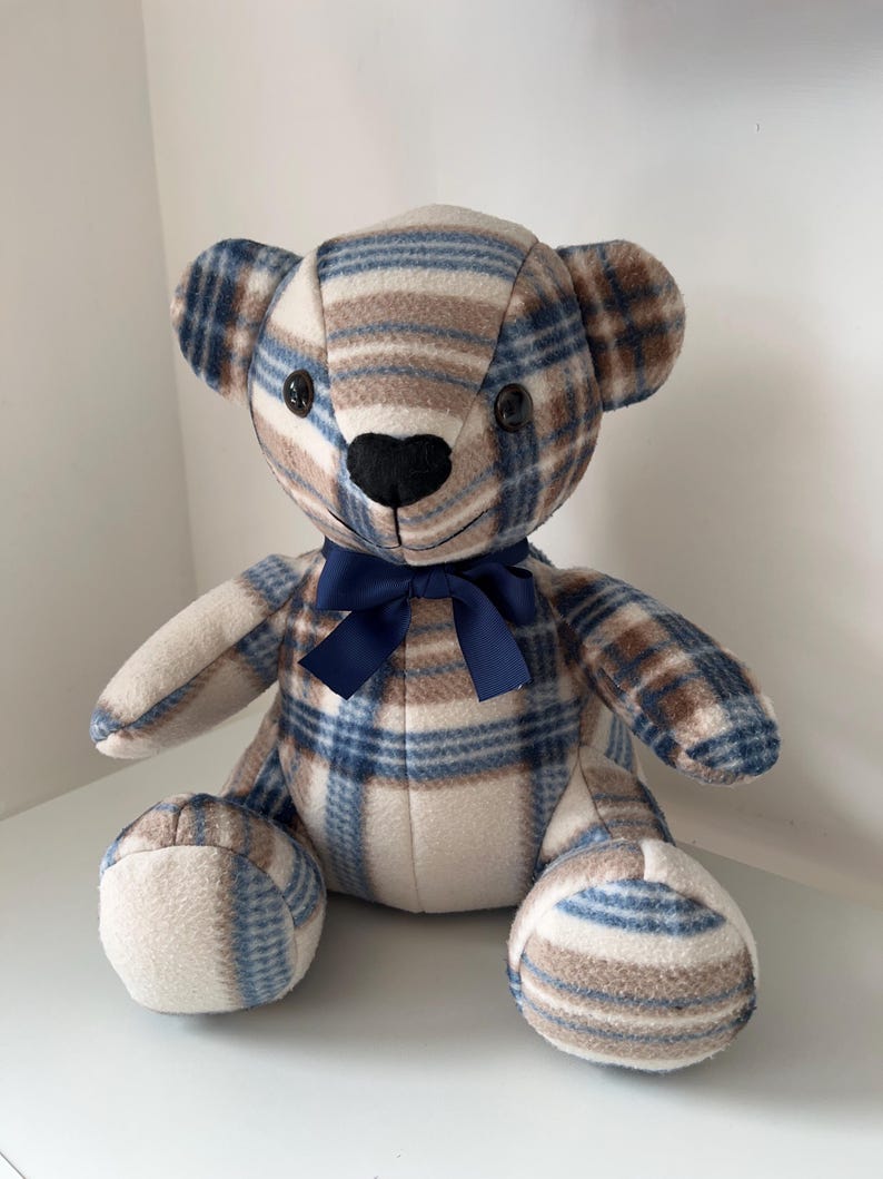 May include: A plush teddy bear with a plaid pattern in shades of blue, brown, and cream. The bear has black button eyes, a black nose, and a navy blue bow. The bear is sitting upright.