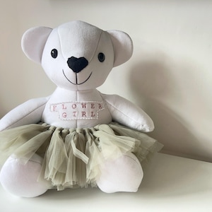 May include: A white teddy bear with a black heart-shaped nose and a stitched smile. The bear wears a light green tulle skirt and a banner that reads "FLOWER GIRL" in pink letters. A cute gift for a flower girl.