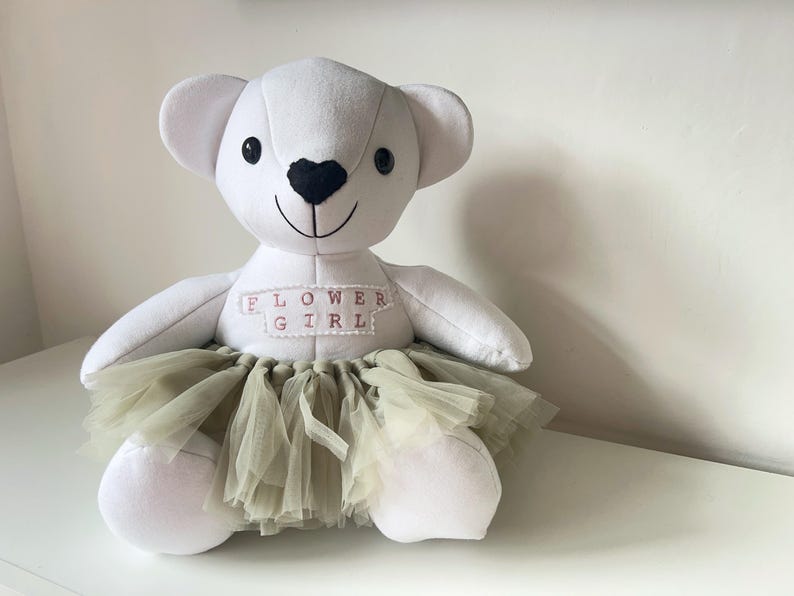 May include: A white teddy bear with a black heart-shaped nose and a stitched smile. The bear wears a light green tulle skirt and a banner that reads "FLOWER GIRL" in pink letters. A cute gift for a flower girl.