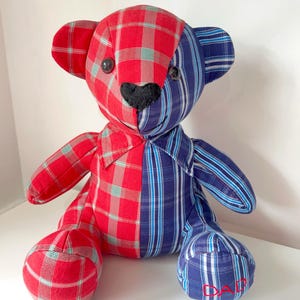May include: A handmade teddy bear with a patchwork design. The bear is split vertically, with one side in red plaid and the other in blue striped fabric. It has black button eyes and a black heart-shaped nose. The word "DAD" is embroidered on one foot.
