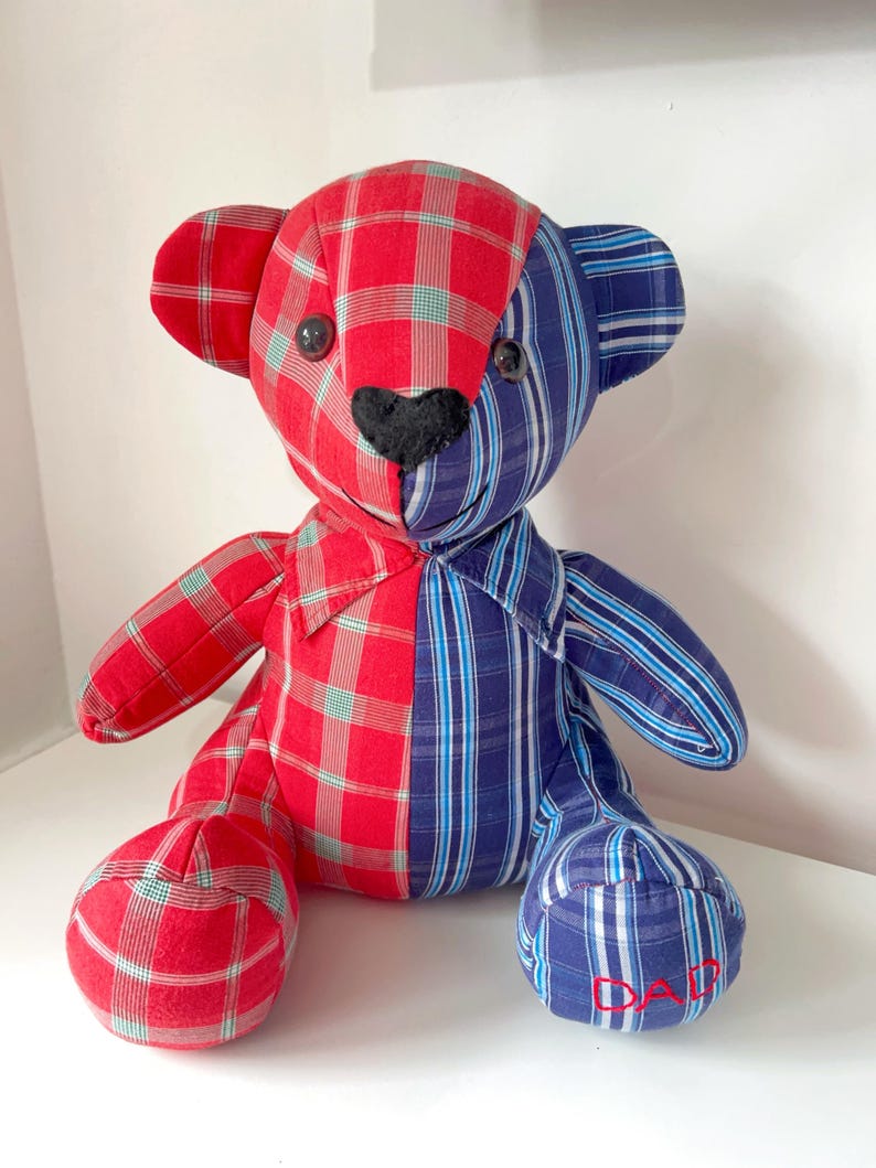 May include: A handmade teddy bear with a patchwork design. The bear is split vertically, with one side in red plaid and the other in blue striped fabric. It has black button eyes and a black heart-shaped nose. The word "DAD" is embroidered on one foot.