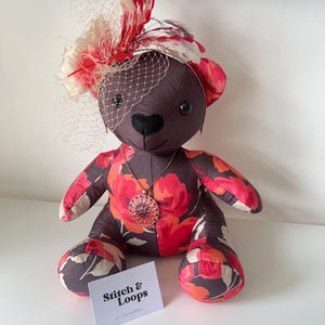 May include: A handmade teddy bear with a floral pattern in red, pink, and orange on a dark brown background. The bear wears a decorative hat with red and white feathers and a veil. A pendant necklace and a business card are also visible.