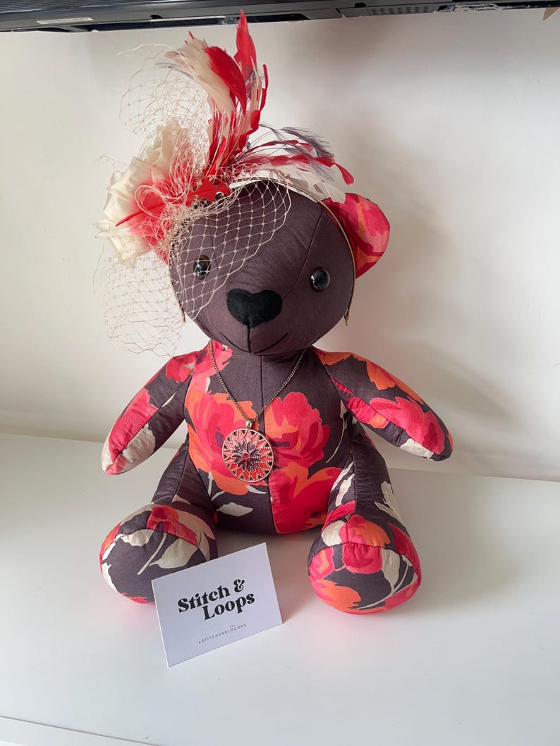 May include: A handmade teddy bear with a floral pattern in red, pink, and orange on a dark brown background. The bear wears a decorative hat with red and white feathers and a veil. A pendant necklace and a business card are also visible.