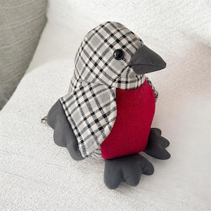 May include: A plush bird toy featuring a head and wings patterned in a grey, black, and cream plaid fabric. The chest is a solid deep red with a herringbone texture. The beak and feet are made of a dark grey fabric, and the eye is a black bead.