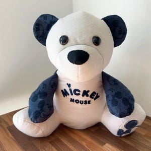 May include: A plush bear toy with a white body, black nose, and dark blue ears and accents. The bear has the words "Mickey Mouse" printed on its chest. The toy is sitting on a wooden surface.