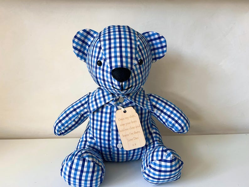 May include: A stuffed teddy bear made of blue and white checkered fabric. The bear has a black nose and eyes, and a small tag with text is attached to its shirt. The bear is sitting upright.