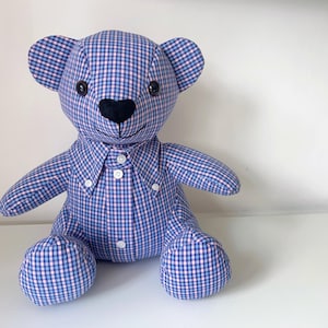 May include: A handmade teddy bear made from blue and pink checkered fabric. The bear has black button eyes, a black heart-shaped nose, and a button-down shirt collar. The bear is sitting on a white surface.