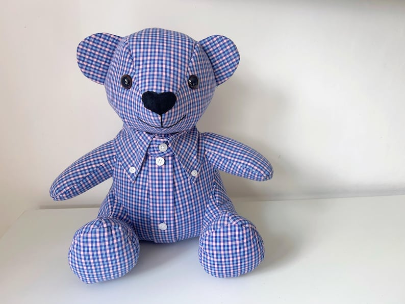 May include: A handmade teddy bear made from blue and pink checkered fabric. The bear has black button eyes, a black heart-shaped nose, and a button-down shirt collar. The bear is sitting on a white surface.