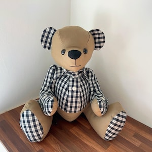 May include: A plush teddy bear with a tan body and black and white checkered accents on the ears, paws, and shirt. The bear has a black nose and button eyes, and is sitting on a wooden surface.