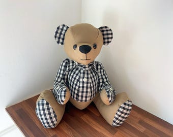 May include: A plush teddy bear with a tan body and black and white checkered accents on the ears, paws, and shirt. The bear has a black nose and button eyes, and is sitting on a wooden surface.