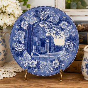 May include: A round ceramic decorative plate featuring a blue and white transferware pattern. The center depicts a historic building in Salem, N.C., with text reading Historic Salem N.C. and South Church Street Moravian Church 1800 Salem College 1772.