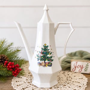 May include: A white ceramic coffee pot with a faceted body and a pointed lid. The front features a colorful illustration of a decorated Christmas tree with a teddy bear and wrapped gifts underneath. The handle and spout are angular in design.