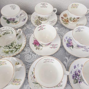 Könnte beinhalten: Eine Sammlung weißer Teetassen und Untertassen, jeweils mit Blumendesigns und dem Namen eines Monats verziert. Die Teetassen haben goldene Ränder und Henkel. Die Untertassen zeigen verschiedene Blumenillustrationen, die eine Vintage-Ästhetik erzeugen.