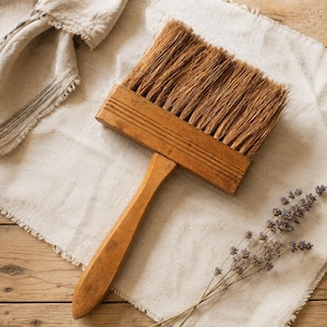 Könnte beinhalten: Ein breiter, flacher Holzbürstenpinsel mit einem langen, stabilen Griff und dicken, groben, hellbraunen Naturborsten. Der rechteckige Holzkopf der Bürste ist auf der Vorderseite mit drei horizontalen Rillen versehen.