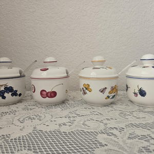 May include: Four white ceramic jars with lids and small spoons. Each jar is decorated with a different fruit or floral design: grapes, cherries, flowers, and berries. The jars are on a white lace tablecloth.