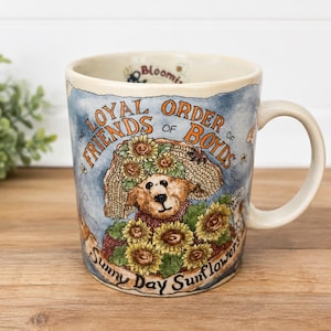 May include: A ceramic mug featuring a brown bear wearing a straw hat surrounded by yellow sunflowers. The text reads The Loyal Order of Friends of Boyds and Sunny Day Sunflowers. The background is a light blue wash with a handle on the right side.