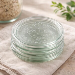 May include: A stack of clear glass jar lids with the embossed word "WECK" and a strawberry design. The circular lids have a slightly raised edge. The lids are stacked on a beige cloth.