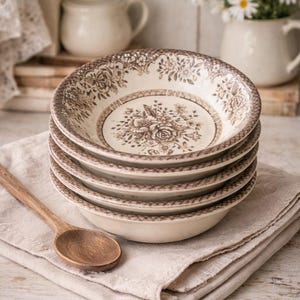 May include: A stack of four cream-colored ceramic bowls with a brown floral pattern. The bowls are stacked on a beige linen napkin, with a wooden spoon to the side. A white vase with daisies is in the background.