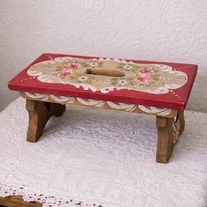 Puede incluir: Un pequeño taburete de madera rectangular con una parte superior roja con un diseño floral. La parte superior tiene un recorte para un asa y está sostenida por cuatro patas marrones. El diseño floral incluye rosas rosas y hojas verdes.