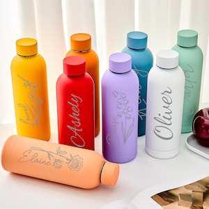 May include: A selection of vibrant, personalised water bottles in various colours: yellow, red, purple, blue, white, and orange. Each bottle features an engraved name and floral design. Some plums are on a white tray.