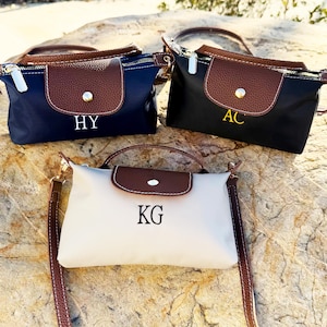 May include: Three personalized handbags in navy blue, black, and off-white, each with a brown flap and handle. The bags feature gold zippers and are embroidered with initials: "HY", "AC", and "KG".