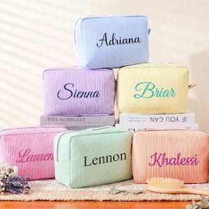 Puede incluir: Bolsas de cosméticos personalizadas en colores pastel, azul, morado, amarillo, rosa, verde y naranja. Cada bolsa tiene un nombre bordado. Están dispuestas sobre una mesa de madera, con un cepillo y una vela.