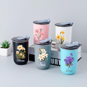 Puede incluir: Una colección de cinco tazas de viaje aisladas con diseños florales. Las tazas son de varios colores, incluyendo negro, rosa, blanco, verde azulado y azul. Cada taza presenta un arreglo floral diferente y un nombre. Las tazas tienen tapas negras con una asa azul claro.