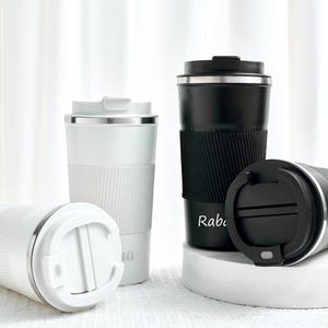Puede incluir: Un conjunto de tazas de viaje personalizadas en blanco y negro. Las tazas blancas tienen los nombres "Erika" y "Anna" impresos. La taza negra tiene el nombre "Rabi" impreso. Cada taza tiene un agarre de silicona acanalado y una tapa plateada.