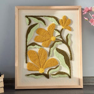 Può includere: Opera d'arte floreale incorniciata. Presenta tre grandi fiori gialli con foglie verdi e marroni. Il design strutturato è racchiuso in una cornice di legno chiaro. Adatta per soggiorno o camera da letto.