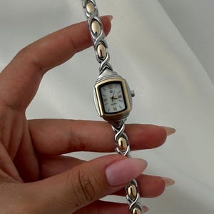 May include: A two-tone wristwatch featuring a rectangular white face with black markers and hands. The band is constructed from alternating silver-tone and gold-tone metal links in an X-shaped pattern, creating a continuous, flexible design.