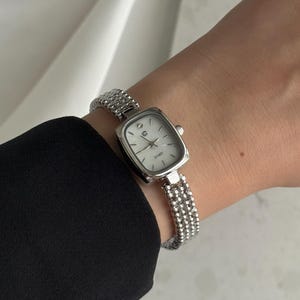 May include: Silver wristwatch with a square face and a beaded strap. The watch features a white dial with black hands and markers. The strap is composed of small, round beads. The watch is worn on a wrist, with a black sleeve visible.