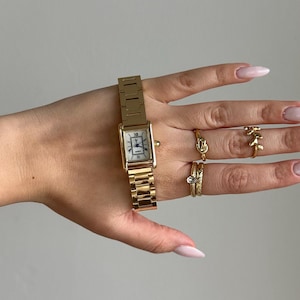 May include: A gold rectangular wristwatch with a white face and gold strap. Several gold rings are on the fingers. The watch is displayed on a hand with long, light pink nails.