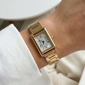May include: A gold-toned rectangular wristwatch with a white face and black Roman numerals. The watch has a gold band and a small blue gem on the side. The watch is on a wrist, and the background is a white surface.