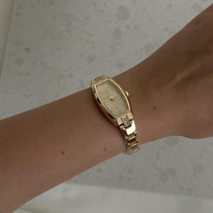 May include: A gold-tone analog wristwatch featuring a tonneau-shaped case with a champagne dial and gold hands. The watch has a link bracelet with small clear stones embedded in the lugs where the strap connects to the watch case.