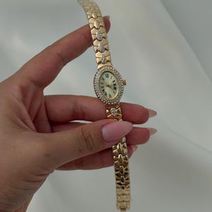 May include: A gold-tone wristwatch featuring an oval-shaped face with Roman numeral markers and a bezel encrusted with small clear stones. The watch band consists of interlocking gold-tone links, with select links accented by small clear stones.