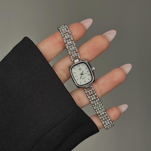 May include: Silver-toned wristwatch with a rectangular face and a beaded band. The watch has a white dial with black hands and markers. The band is made of small, round beads. The watch is a classic accessory.
