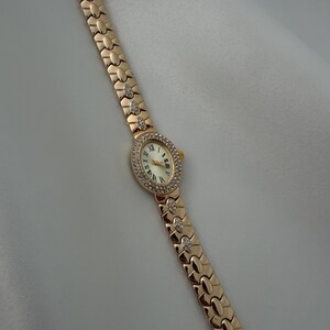 May include: A gold-tone wristwatch featuring an oval-shaped face with a white dial and black Roman numeral markers. The bezel is encrusted with small clear stones. The metal link bracelet includes decorative segments accented with clear stones.