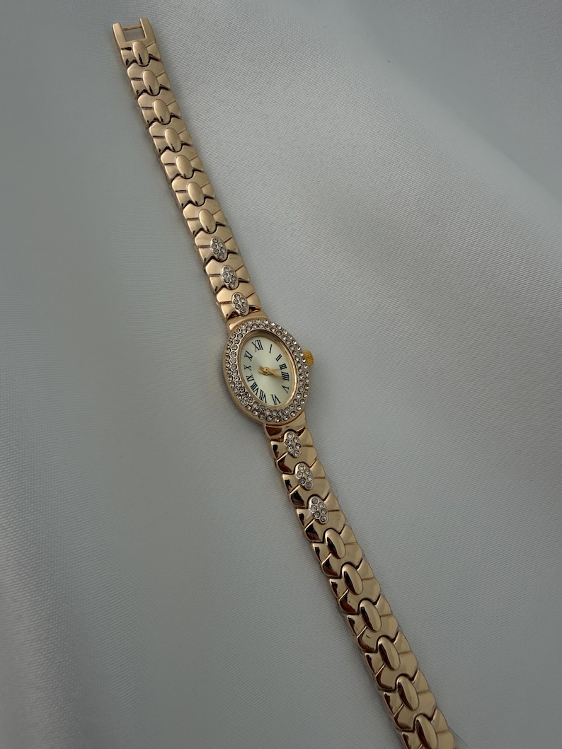 May include: A gold-tone wristwatch featuring an oval-shaped face with a white dial and black Roman numeral markers. The bezel is encrusted with small clear stones. The metal link bracelet includes decorative segments accented with clear stones.