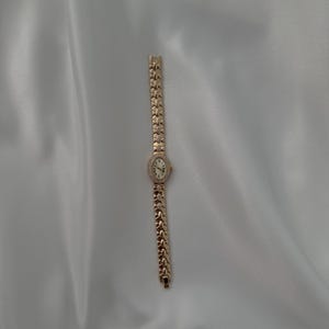 May include: A gold-tone wristwatch featuring an oval-shaped face with a white dial and a bezel accented with small clear stones. The watch has a decorative link bracelet composed of repeating X-shaped metal segments with small embedded stones.