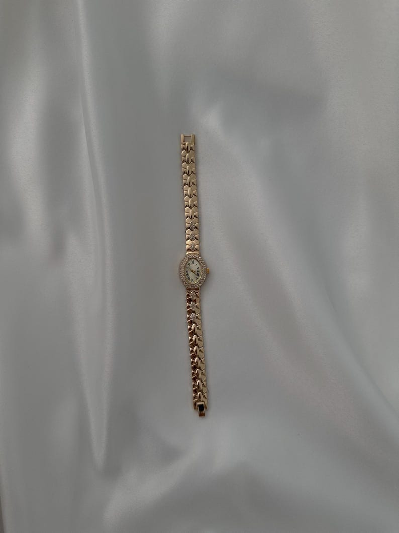 May include: A gold-tone wristwatch featuring an oval-shaped face with a white dial and a bezel accented with small clear stones. The watch has a decorative link bracelet composed of repeating X-shaped metal segments with small embedded stones.