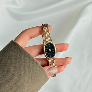 Puede incluir: Un reloj de pulsera dorado con una esfera ovalada negra y n&uacute;meros romanos. El reloj tiene una correa de eslabones dorados y se sostiene en una mano. La esfera del reloj dice "QUARTZ".
