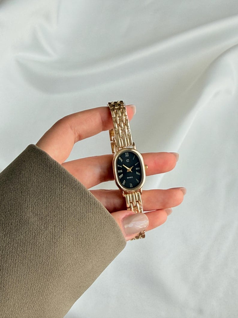 Puede incluir: Un reloj de pulsera dorado con una esfera ovalada negra y n&uacute;meros romanos. El reloj tiene una correa de eslabones dorados y se sostiene en una mano. La esfera del reloj dice "QUARTZ".