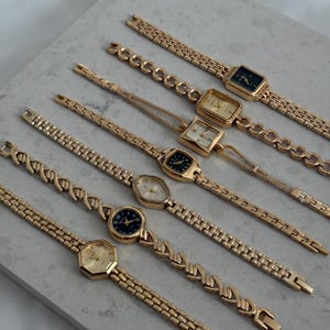 May include: A collection of gold-toned wristwatches with various face shapes and band designs. The watches feature rectangular, square, and oval faces with gold or black dials. The bands include chain-link and patterned styles.