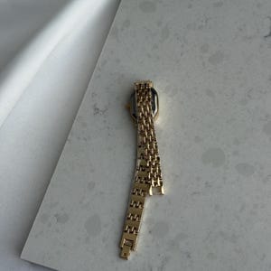 Puede incluir: Un reloj de pulsera dorado con una esfera rectangular y una correa de eslabones. El reloj se muestra sobre una superficie gris claro, con tela blanca en el fondo. La correa del reloj est&aacute; parcialmente desabrochada.