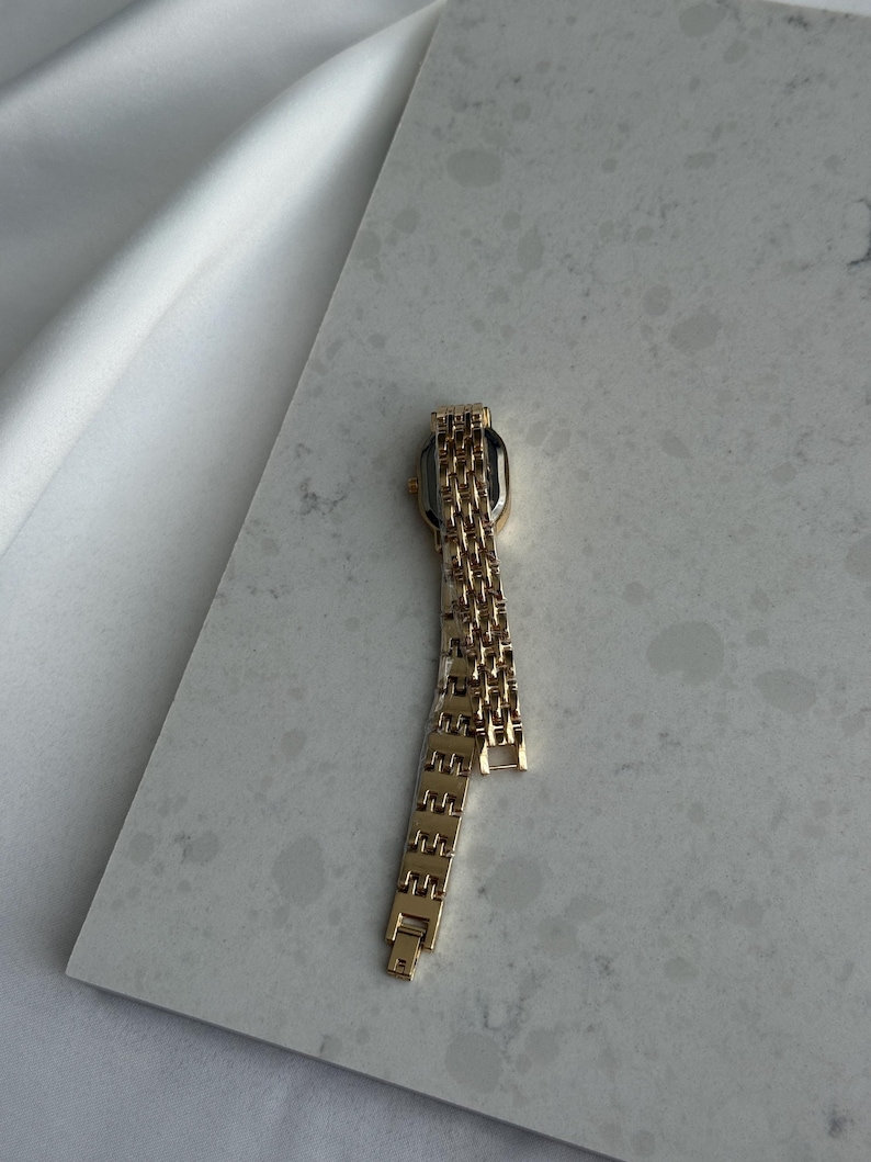 Puede incluir: Un reloj de pulsera dorado con una esfera rectangular y una correa de eslabones. El reloj se muestra sobre una superficie gris claro, con tela blanca en el fondo. La correa del reloj est&aacute; parcialmente desabrochada.