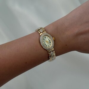 May include: A gold-tone oval wristwatch featuring a bezel encrusted with clear crystals. The watch face has a cream background with black Roman numeral hour markers and gold hands. The metal link bracelet is also adorned with clear crystals.