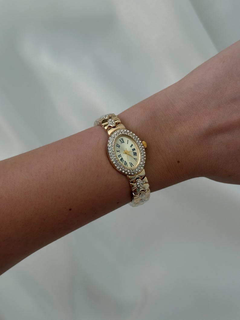May include: A gold-tone oval wristwatch featuring a bezel encrusted with clear crystals. The watch face has a cream background with black Roman numeral hour markers and gold hands. The metal link bracelet is also adorned with clear crystals.